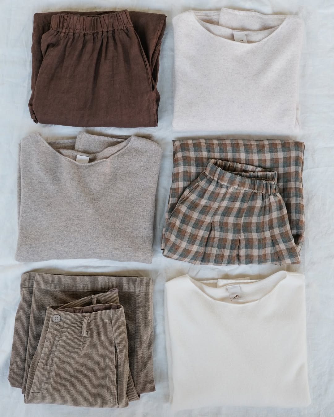 Folded Montaigne linen and wool clothing items including a brown sweater, plaid culottes, and beige pants on a white surface.